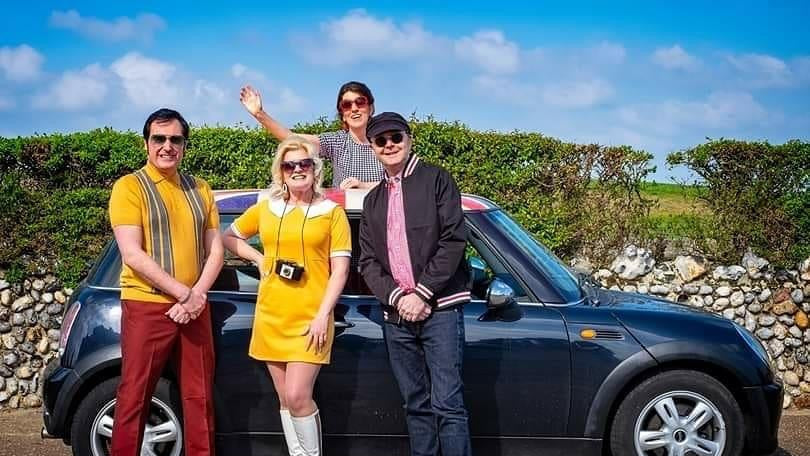 Four people posing in front of a black car with a scenic background